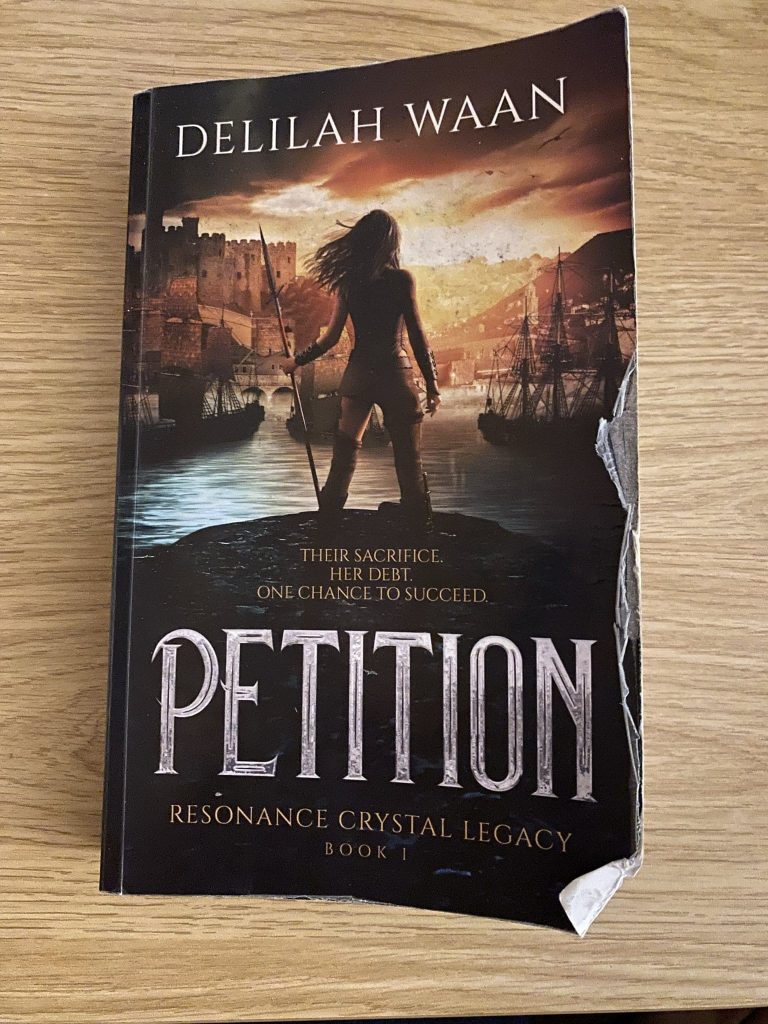Photo of a battered print-on-demand paperback edition of Petition by Delilah Waan, showing visible peeling of the matte lamination on the front cover.